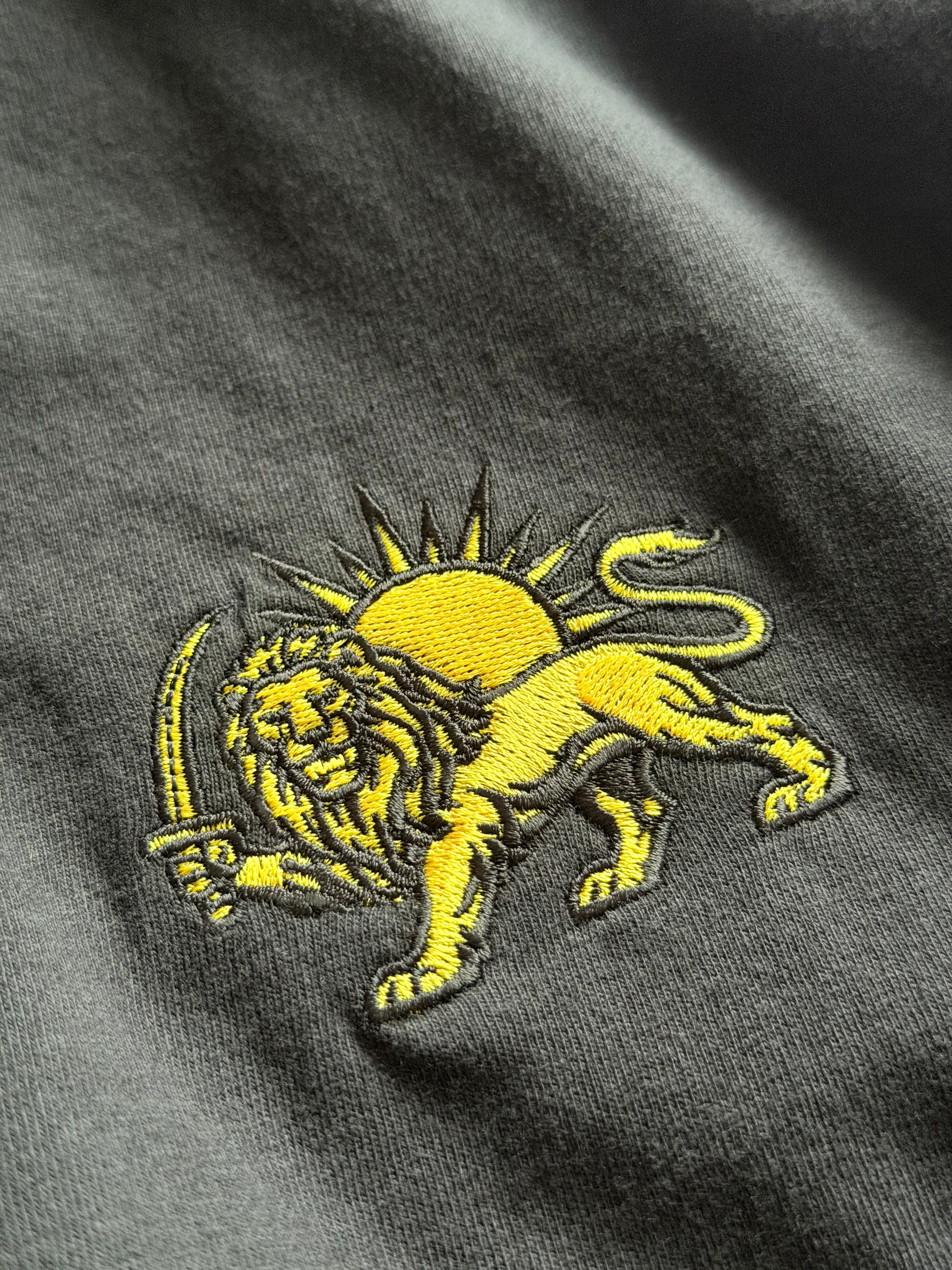 Close-up of a dark gray fabric featuring a yellow embroidered lion holding a sword with a yellow sun behind it.